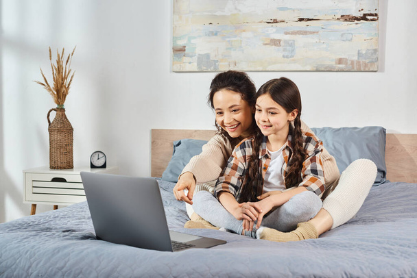 mãe e filha sentam-se em uma cama, absorvidos em uma tela de laptop, engajando-se em tempo de qualidade juntos em casa. - Foto, Imagem