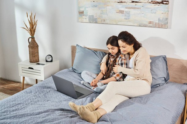 Mother and daughter sitting on a bed, engrossed in a laptop screen, sharing a special moment together. - Foto, afbeelding