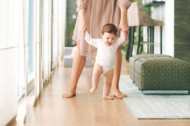 Мама помогает очаровательному ребенку сделать свои первые шаги, держа ребенка за обе руки - Фото, изображение
