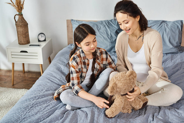 A mother and daughter sit together on a bed, deep in conversation while holding a teddy bear. - Foto, Imagen