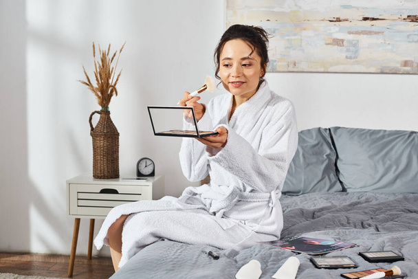 Une femme brune en peignoir blanc s'assoit sur un lit tenant une ombre à paupières, entourée de cosmétiques, se maquillant le matin. - Photo, image