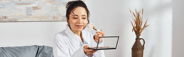 A brunette woman in a white robe holding eye shadows palette in a cozy setting. - Photo, Image