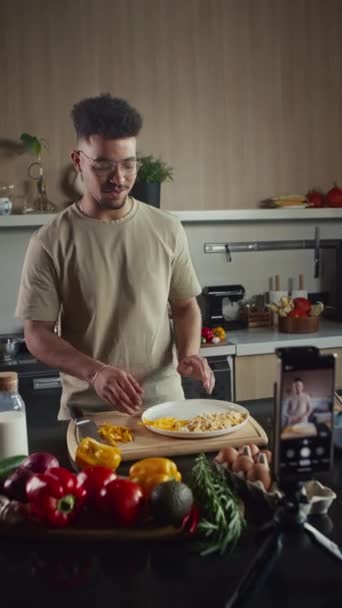 Pionowe ujęcie kulinarnego blogera serwującego danie na talerzu i rozmawiającego przez kamerę telefoniczną podczas filmowania jedzenia vlog w kuchni - Materiał filmowy, wideo