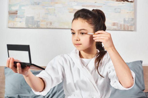A brunette preteen in a white bath robe sits on a bed, applying makeup to her face with different cosmetics. - Photo, Image