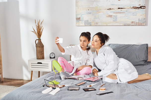 Moeder en dochter in witte badjassen zitten sereen samen op het bed en delen een vredig moment van verbondenheid. - Foto, afbeelding