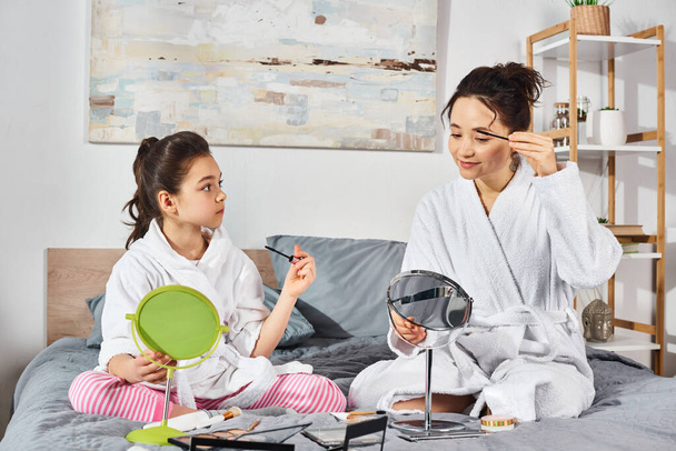 Брюнетка мать и дочь в белых халатах сидят вместе на уютной кровати, разделяя нежный момент, нанося макияж - Фото, изображение