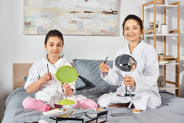 Спокойная сцена, где брюнетка мать и дочь в белых халатах удобно сидят на кровати, нанося макияж - Фото, изображение