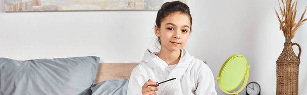 Ein Mädchen mit brünetten Haaren sitzt in einem weißen Bademantel auf einem Bett und hält eine Wimperntusche in der Hand. - Foto, Bild
