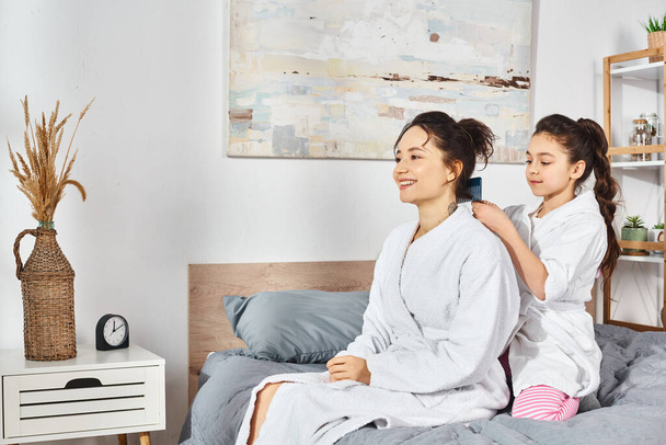 Δύο γυναίκες, μια μητέρα και η κόρη, και οι δύο με καστανά μαλλιά, κάθονται σε ένα κρεβάτι με άσπρες ρόμπες, μοιράζονται μια ειρηνική στιγμή μαζί. - Φωτογραφία, εικόνα