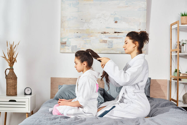 Brunette mère et fille en peignoirs blancs assis sur le lit, partageant un moment d'amour pendant que la mère brosse les cheveux des filles. - Photo, image