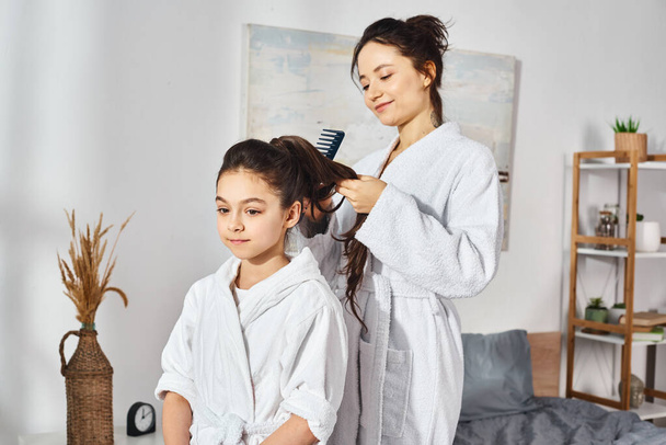Eine brünette Mutter frisierte ihren Töchtern die Haare auf einem Bett, beide trugen weiße Bademäntel. - Foto, Bild