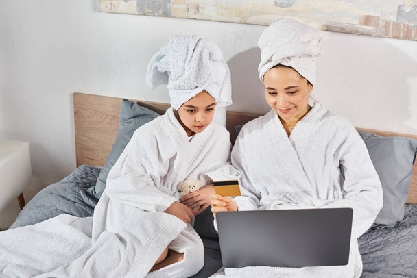 Zwei Frauen, eine brünette Mutter und ihre Tochter, in weißen Bademänteln auf einem Bett sitzend, mit einem Laptop beschäftigt. - Foto, Bild