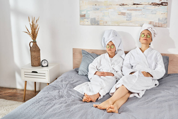 Брюнетка мати і дочка в білих халатах для ванни, сидячи на ліжку, з рушниками, загорнуті навколо їх голови. - Фото, зображення