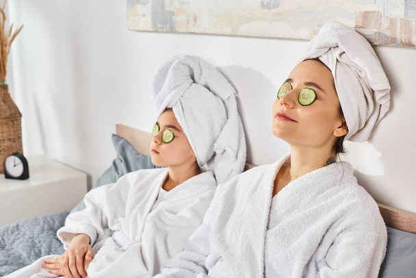 Dwie brunetki w białych szatach kąpielowych cieszące się zabiegiem spa z plastrami ogórkowymi na oczach. - Zdjęcie, obraz