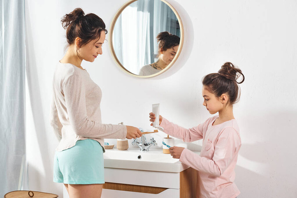 A brunette woman and her preteen daughter stand together in front of a sink in a modern bathroom, engaging in beauty and hygiene routines. - Foto, Imagem