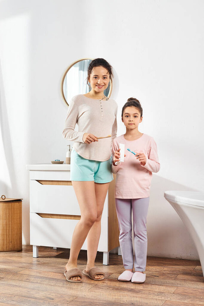 A brunette woman stands with her preteen daughter in a modern bathroom, engaging in beauty and hygiene routines together. - 写真・画像