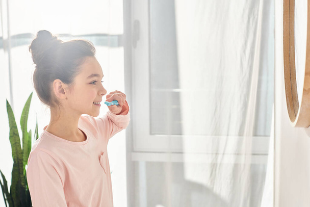 Μια μελαχρινή κοπέλα πριν την εφηβεία ασχολείται με την ομορφιά και την υγιεινή το πρωί, βουρτσίζοντας τα δόντια μπροστά από έναν καθρέφτη. - Φωτογραφία, εικόνα