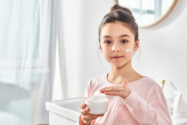A young girl with dark hair holds a jar of cream in her hands, showcasing a skincare or beauty routine in a modern bathroom. - Foto, afbeelding