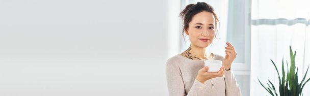 Eine brünette Frau hält ein Glas Creme in der Hand und blickt mit einem gelassenen Gesichtsausdruck direkt in die Kamera. - Foto, Bild