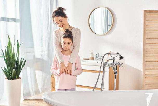 Een vrouw met brunette haar en haar tiener dochter staan in een moderne badkamer, bezig met hun schoonheid en hygiëne routine. - Foto, afbeelding