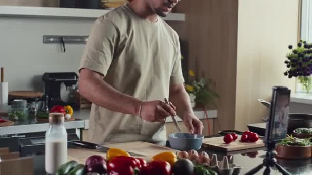 Food blogger χτυπώντας συστατικά σε μπολ και λέγοντας πώς να μαγειρέψουν το πρωινό, ενώ μαγνητοσκόπηση συνταγή βίντεο με κινητό τηλέφωνο στο τρίποδο στην κουζίνα - Πλάνα, βίντεο