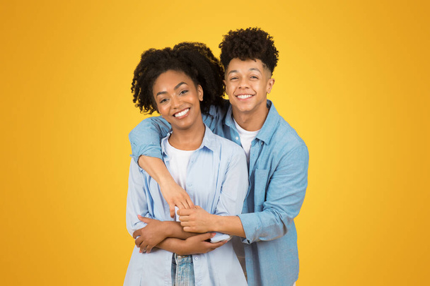 カジュアルなブルーシャツの輝かしい幸せな若い女性と男性は暖かく抱擁し,明るい黄色の背景に彼女の周りのマンションアームと本物の笑顔を共有しています. 愛,関係,広告,そして提供 - 写真・画像