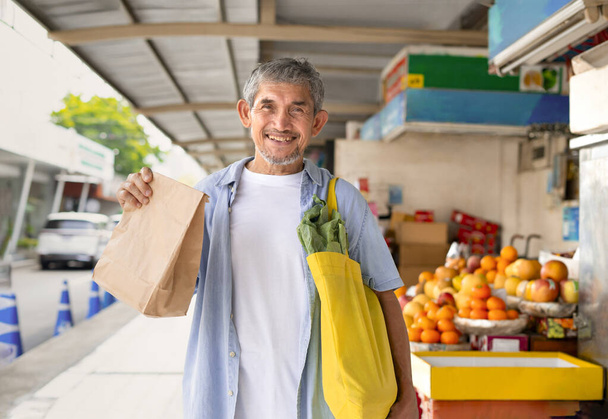 portrait senior man holding a yellow eco-friendly cloth bags to put vegetables and paper bag, background the community fruits market. concept of plastic free, eco freindly - Photo, Image