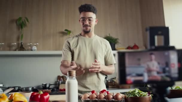 Rack focus shot van jonge mannelijke voedsel blogger vertellen over ingrediënten op smartphone camera gehouden door statief tijdens het filmen koken show in de keuken - Video