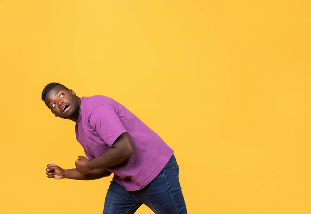Pelokas musta mies violetti t-paita kumartuu puolustava poseeraa ja katselee ylös, odottaa vaaraa ylhäältä, poseeraa vastaan keltainen studio tausta. Vapaa tila tekstille - Valokuva, kuva