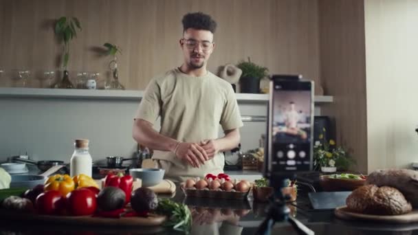 キッチンでビデオレシピを撮影しながら三脚が保有する携帯電話の前で話す食材を示す若いフードブロガーのラックフォーカスショット - 映像、動画