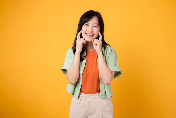 hygiène dentaire avec une charmante jeune femme asiatique des années 30, élégamment vêtue d'une chemise orange et d'un pull vert pointe ses dents sur un fond jaune, soulignant l'importance des soins dentaires. - Photo, image