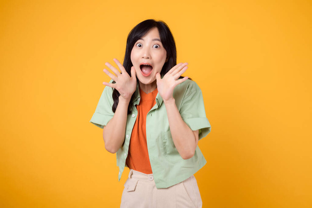 Energetic young Asian woman 30s wearing a green and orange shirt passionately shouting with excitement. Isolated on a yellow background, representing the concept of discount shopping promotion. - Photo, Image