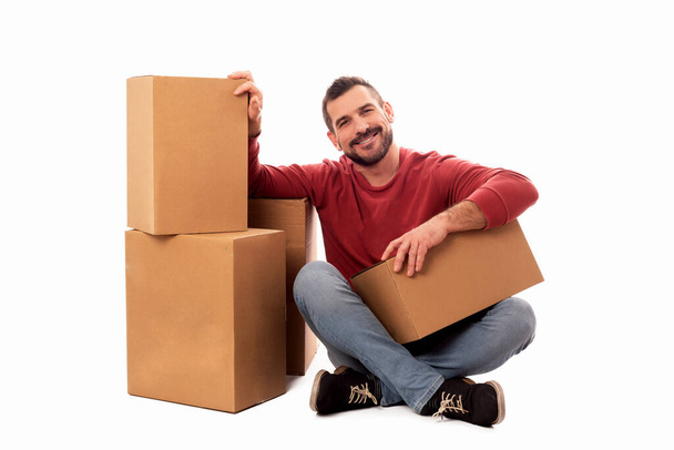 Smiling seated man with beard and red sweater on white background surrounded by large cardboard boxes. Home delivery service. Moving help service. Free shipping - Photo, Image