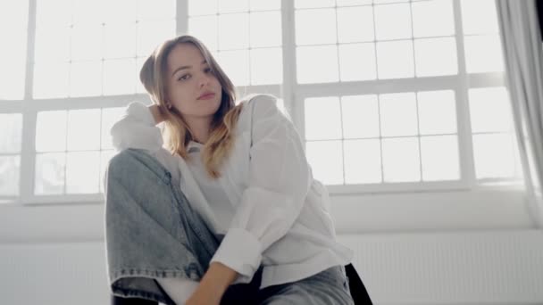 Стильная женщина в джинсах и белой рубашке сидит и позирует на камеру во время съемок в студии. Привлекательная модель в повседневной одежде, смотрящая в камеру - Кадры, видео