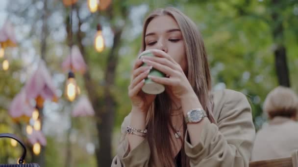 Jonge mooie vrouw zit in een café op straat en drinkt verse koffie. Vrouwelijke hebben pauze en drinken warme thee in het park buiten - Video