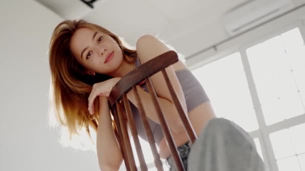 Gyönyörű fiatal nő ül a széken, és pózol a stúdióban. Elbűvölő női modell mosoly és a kamerába néz - Felvétel, videó