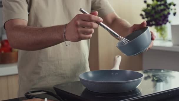 Обрізаний знімок невпізнаваного чоловіка, який поливає збиті яйця з миски на сковороду на плиті, готуючи сніданок на кухні вдома - Кадри, відео