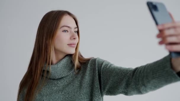 Σίγουρη ελκυστική γυναίκα σε ζεστό πουλόβερ κρατήσει το κινητό τηλέφωνο στο χέρι και να κάνει selfie σε λευκό φόντο. Νεαρή όμορφη γυναίκα θέτουν και να λάβει αυτο εικόνα στο smartphone. - Πλάνα, βίντεο