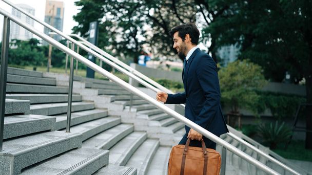 Skilled business man walking up stairs at park or city while holding bag in the hand Professional project manager going to workplace. Represent growth, getting promotion, increasing skill. Exultant. - Photo, Image
