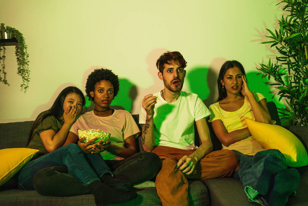 Barátok a kanapén sokkos arccal, miközben egy horrorfilmet néznek, zöld fény kísérteties hangulatot teremt.. - Fotó, kép