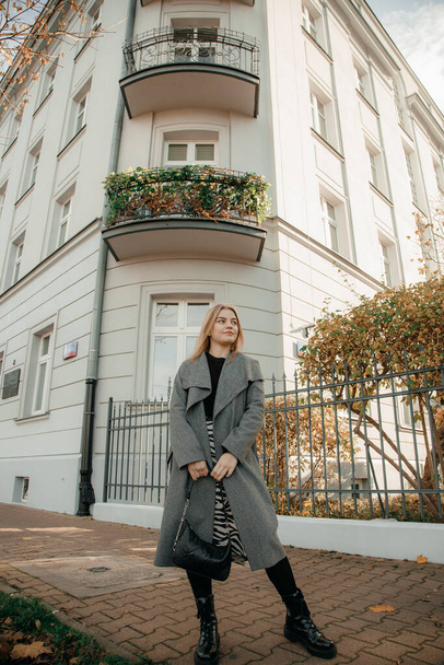 Portret van een stijlvolle glimlachende jonge vrouw tegen de achtergrond van de bouw in de stad. Modieuze blonde vrouw draagt een grijze jas en een zwarte handtas. Jonge vrouw toerist wandelen de in de oude stad. - Foto, afbeelding