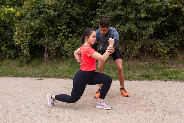 twee personen die deelnemen aan een stretching oefening buiten op een grindpad, omgeven door groen. Een individu, gekleed in zwarte legging en een oranje top, is in een longende positie. - Foto, afbeelding