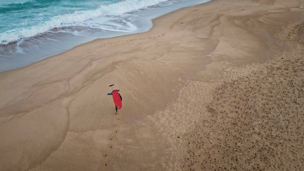 Вид с воздуха серфер прогуливаясь по песчаному пляжу оставляя следы. Серфбордист, несущий доску, шагая мокрым песком с пенной океанской водой по живописной морской природе замедленного хода. Концепция активного досуга - Фото, изображение