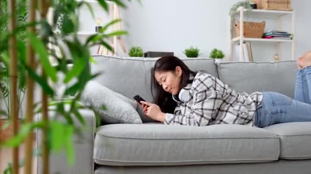 Egy fiatal nő telefonál a kanapén. Ázsiai tinédzser csevegés a szociális média, miközben fekszik a kanapén. - Felvétel, videó