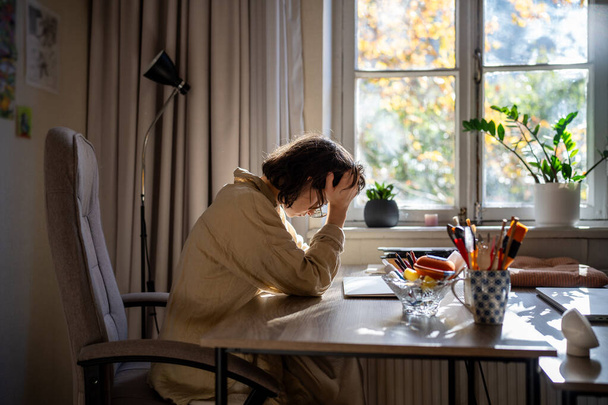 Κουρασμένη εξαντλημένη γυναίκα κάθεται στο τραπέζι κρατώντας το κεφάλι νωρίς το πρωί μετά τη δουλειά όλη τη νύχτα. Κορίτσι εργασιομανής έχει πονοκέφαλο αισθάνεται άσχημα κάθεται στο χώρο εργασίας γραφείο στο σπίτι. Απόμακρη απομακρυσμένη εργασία. - Φωτογραφία, εικόνα