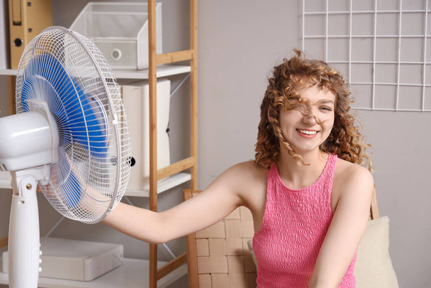 Krásná šťastná mladá žena s elektrickým ventilátorem sedí na židli v obývacím pokoji - Fotografie, Obrázek