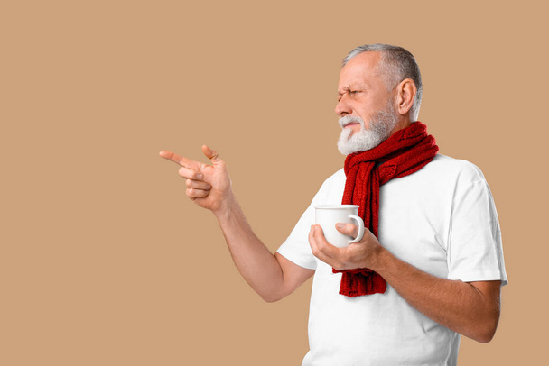 Alter Mann mit Halsschmerzen hält eine Tasse Tee in der Hand und zeigt auf etwas auf beigem Hintergrund - Foto, Bild