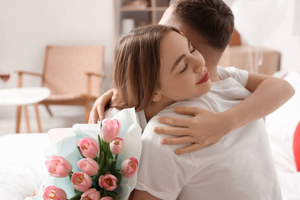 Gyönyörű fiatal pár csokor virágokkal ölelkezve és ünnepelve Valentin-napot otthon - Fotó, kép