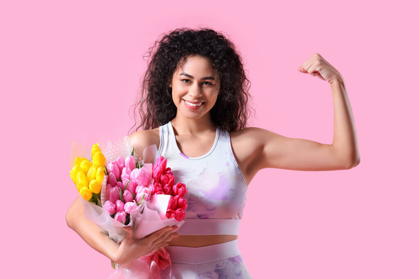 Jeune femme afro-américaine en tenue de sport avec différents bouquets de fleurs montrant des muscles sur fond rose. Célébration de la Journée internationale de la femme - Photo, image