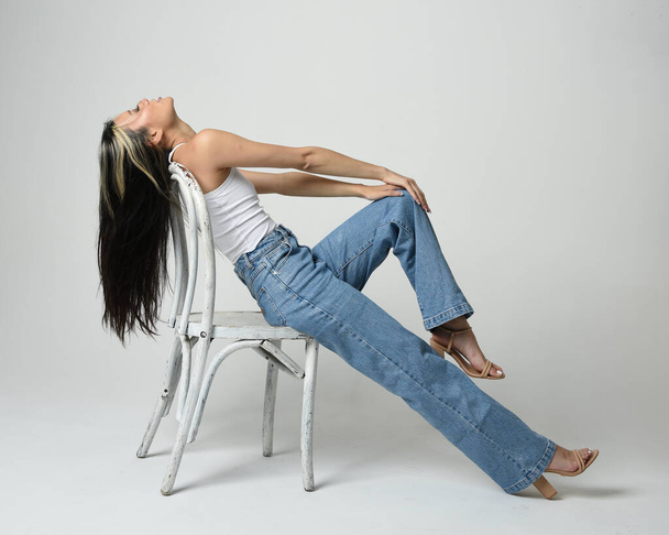 Full length portrait of brunette female asian model wearing casual clothes, white singlet shirt, denim jean pants. Comfortable seated pose on wooden chair. Isolated on white studio background. - Photo, Image